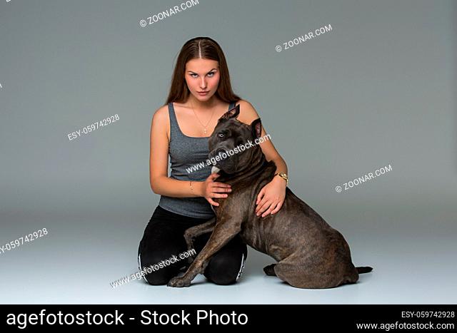 Beautiful sporty young woman sitting on floor and hugging adult grey amstafford terrier dog. Studio shot over gray background. Copy space