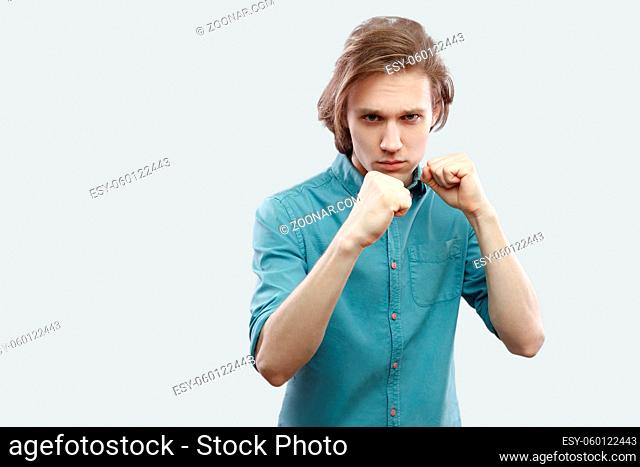 Portrait of serious handsome long haired blonde young man in blue casual shirt standing in boxing fists pose and looking at camera
