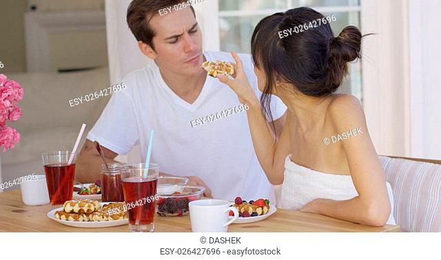 Woman wrapped in towel trying to feed annoyed man a waffle during breakfast time at wooden table. Includes copy space