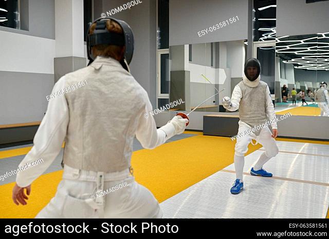 Two swordsmen wearing uniform and protective helmet fighting duel during training in sport gym room. Sportsmen in fencing club
