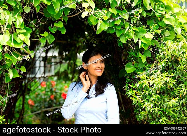 Closeup portrait of confident smiling happy pretty young woman in blue shirt enjoying a fresh new day, isolated background of green shrubs and flowers