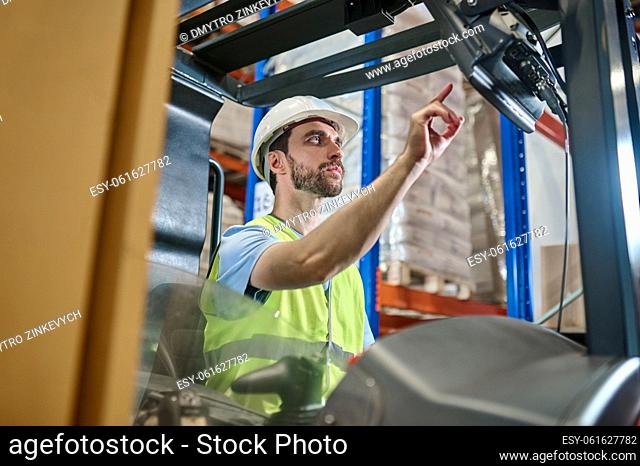 Serious focused young warehouse employee in a reflective vest and helmet starting the lift truck