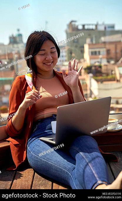 Smiling woman waving hand to video call on laptop while sitting at roof terrace