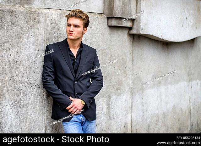 Handsome blond young man, blue eyes, leaning against white, rough stone wall, looking up to a side