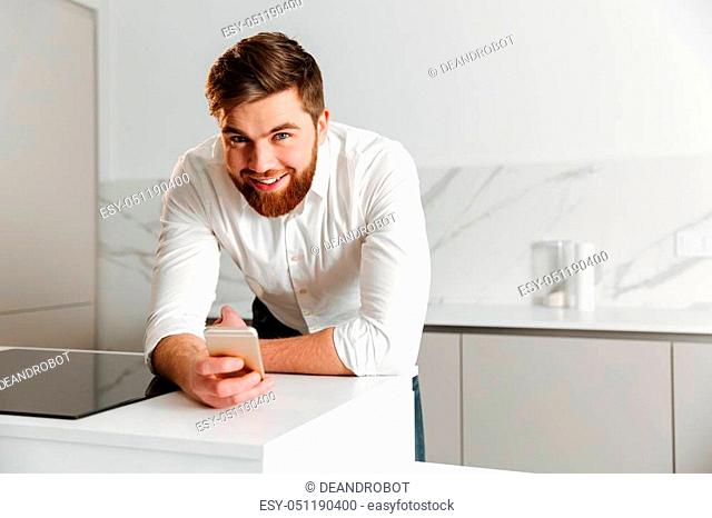 Portrait of a smiling young businessman dressed in white shirt holding mobile phone while leaning on a table indoors