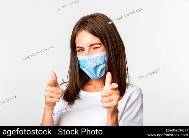 Covid-19, health and social distancing concept. Sassy smiling brunette girl in medical mask winking flirty at camera and pointing fingers