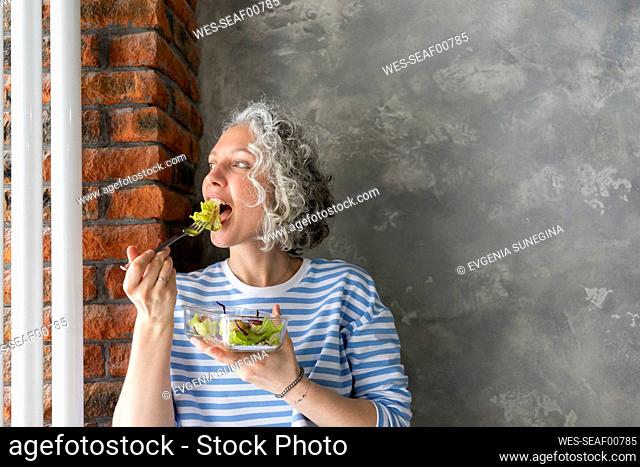 Woman eating fresh salad for lunch in front of gray wall