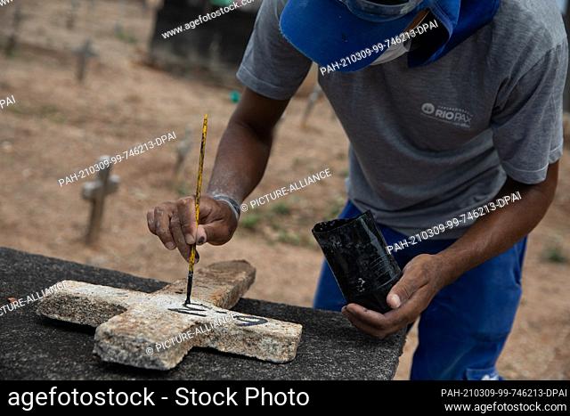 06 March 2021, Brazil, Rio De Janeiro: Douglas Silva, who works at Iraja Cemetery, writes the date on a cross before a burial