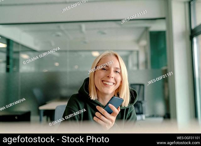 Happy blond businesswoman holding smart phone in office