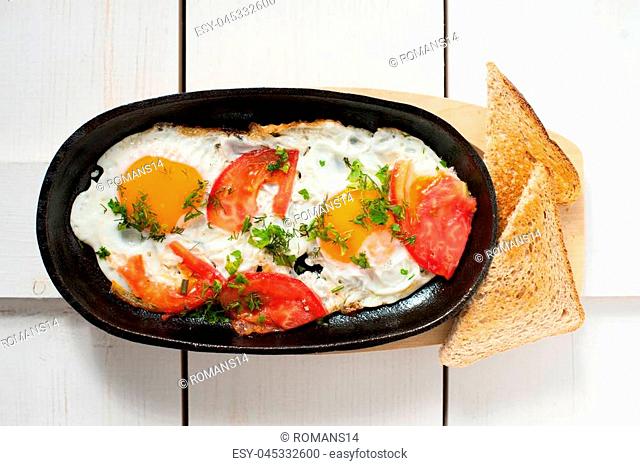 Scrambled eggs with fresh tomatoes and fresh herbs fried in a pan. Served with bread toast. Placed on a white wooden Board