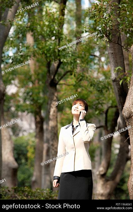 Businesswoman talking on smart phone below trees in park