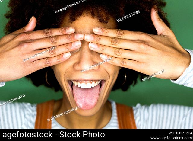 Cheerful young woman sticking out tongue while covering eyes against green wall