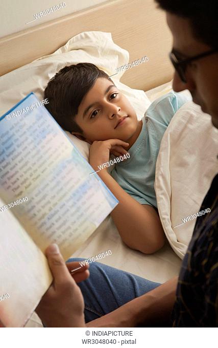 Boy relaxing on the bed while his father is reading him a story
