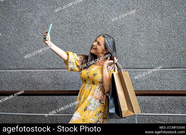 Happy woman taking selfie with shopping bags in front of gray wall