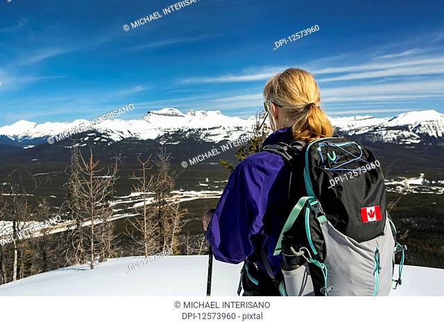 Female hiker on snow-covered pathway with snow-covered mountains, blue sky and clouds in the background; Lake Louise, Alberta, Canada