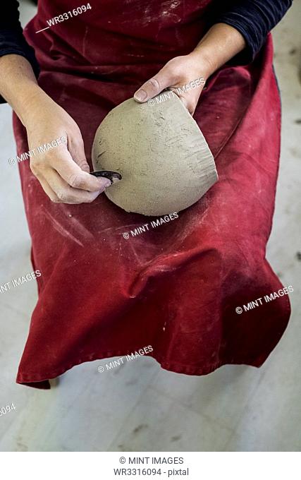 High angle close up of ceramic artist wearing red apron sitting in her workshop, working on clay vase