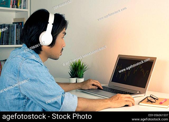 Asian Forex Trader or Investor or Businessman Wear Jean Shirt and Headphone Looking Forex or Stock Chart in Laptop. Casual Businessman Working From Home by...