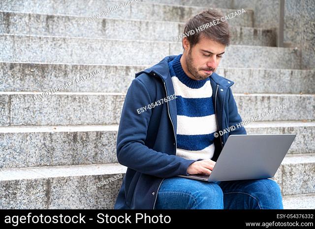 young serious expression, man freelancer working with laptop outdoors sitting at stair steps, in urban scenery