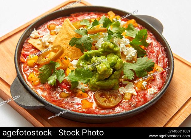 Homemade fresh traditional dish Shakshuka from fried eggs, tomatoes, pepper, vegetables and herbs in a pan on a wooden board on a light grey background