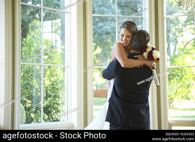 Bride and groom in an embrace, groom lifting up bride standing by large bank of windows
