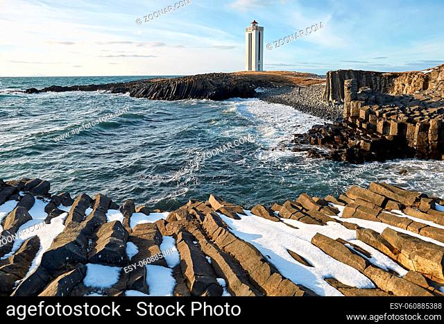 Light lighthouse on the promontory in Iceland on the background of the blue sky with clouds. In front of it there is a rocky shore with remains of snow and a...