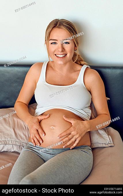 Happy smiling attractive young pregnant woman cradling her bare baby bump in her hands as she relaxes on a bed looking up at the camera