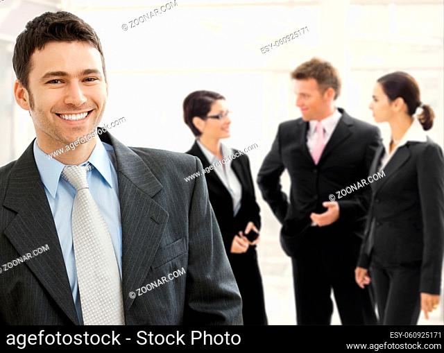 Business team standing in office lobby. Happy businessman in front, smiling and looking at camera, others talking in the background