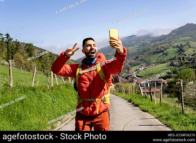 Happy man gesturing peace sign taking selfie through smart phone on sunny day