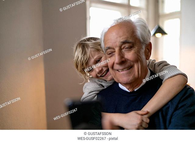 Happy grandson hugging grandfather with cell phone at home