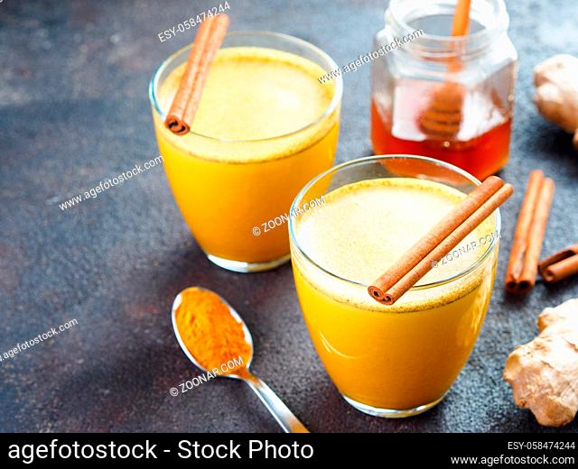 Healthy drink golden turmeric latte in glass.Gold milk with turmeric, ginger root, cinnamon sticks, turmeric powder and honey over black cement background