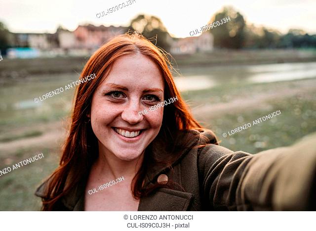 Young woman with long red hair smiling for selfie on riverside, personal perspective, Florence, Tuscany, Italy