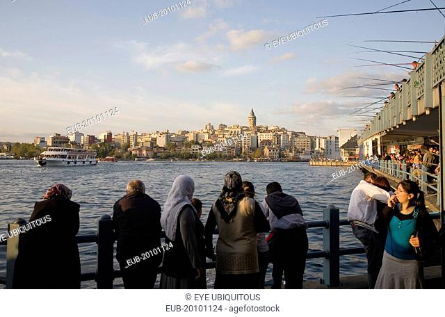 Sultanahmet. Crowd leaning on railings beside the Bosphorus admiring view to Galata tower with rods of men fishing from Galata bridge extended out above