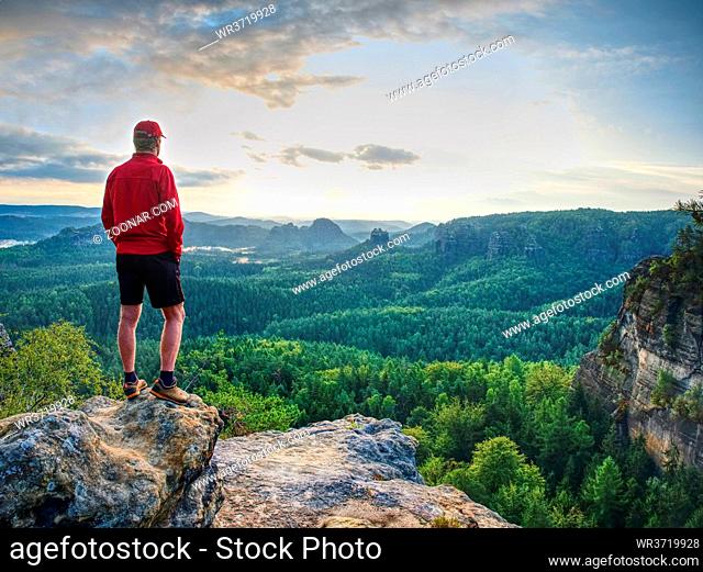 Fit mountain climber or hiker on a rocky summit looking down onto forest in a green valley against Sun at horizon