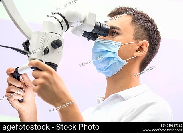 Cute doctor looks into the dental microscope which he holds with his hands on the light background. He wears a white uniform and a blue medical mask