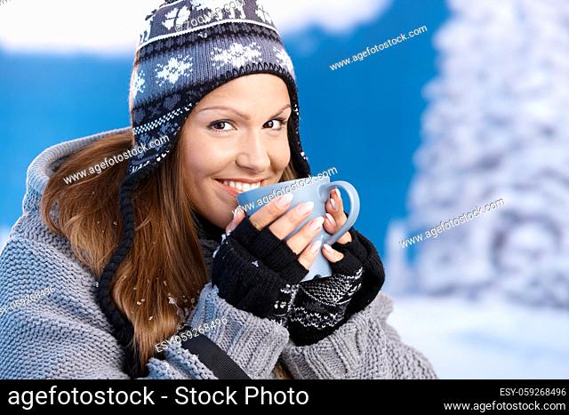 Attractive young female dressed up warm for skiing wearing cap and gloves drinking hot tea smiling front of winter landscape