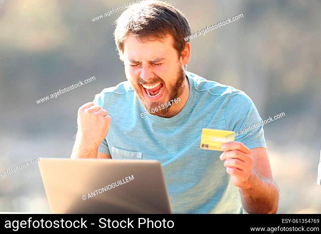 Excited man holding credit card buying online on laptop celebrating success outdoors