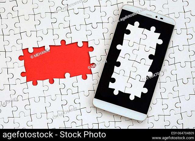 A modern big smartphone with several puzzle elements on the touch screen lies on a white jigsaw puzzle in an assembled state with missing elements