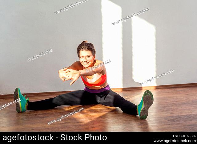 Beginner stretching, warm-up. Positive woman with bun hairstyle and in sportswear doing easy stretches and smiling at camera, practicing at home