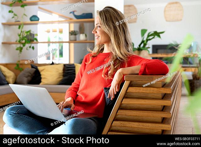 Smiling blond woman sitting with laptop on couch in living room while looking away