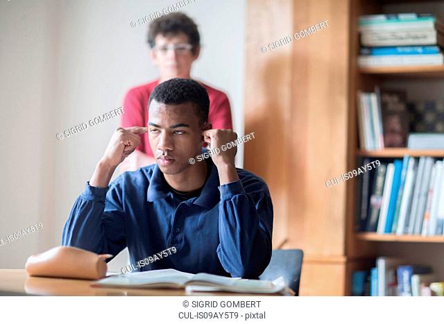 Young male high school student sitting at desk with fingers in ears and teacher standing behind