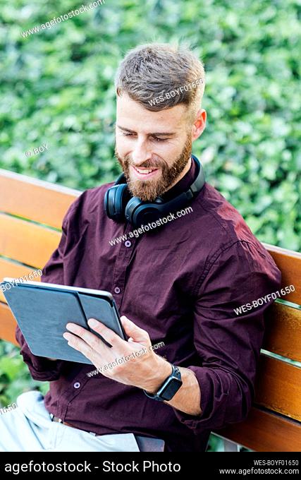 Smiling businessman with headphones using digital tablet while sitting on bench