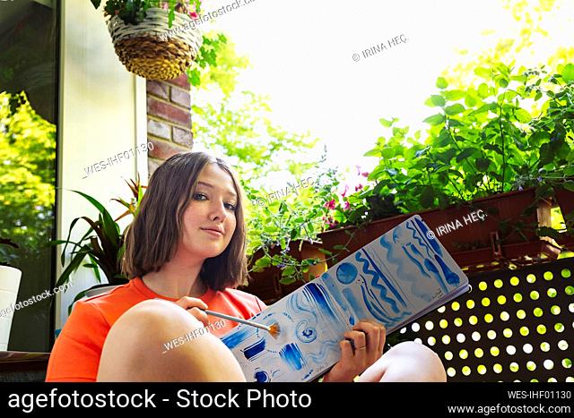 Teenage girl showing watercolor painting on sketch pad sitting on balcony