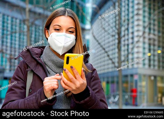 Portrait of a young woman with medical face mask checking notifications on smartphone on urban background. Copy space