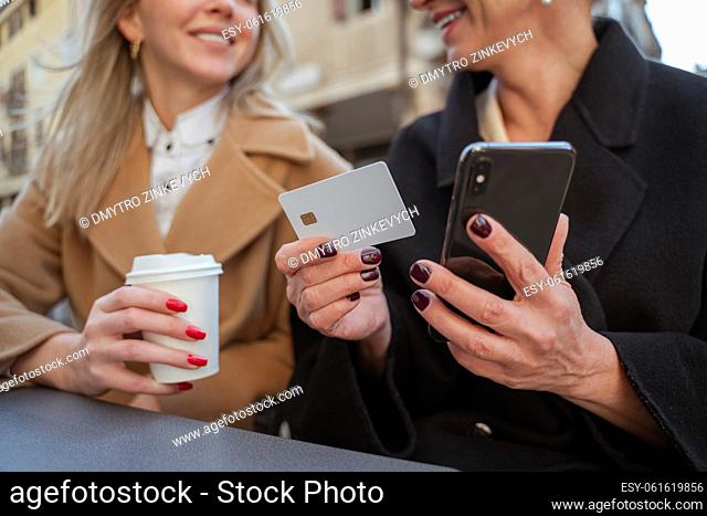 Cropped photo of a woman with a credit card and smartphone sitting at a cafe table beside a smiling blonde