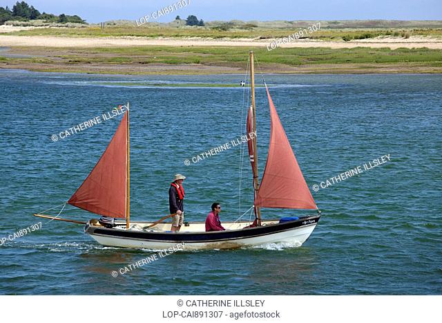 England, Norfolk, Wells Next The Sea. Two men sailing a boat down the channel between the harbour and beach at Wells Next The Sea
