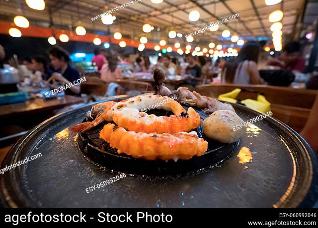 Seafood on steaming and grilling cooker pot over blurred view of people sitting in restaurant background