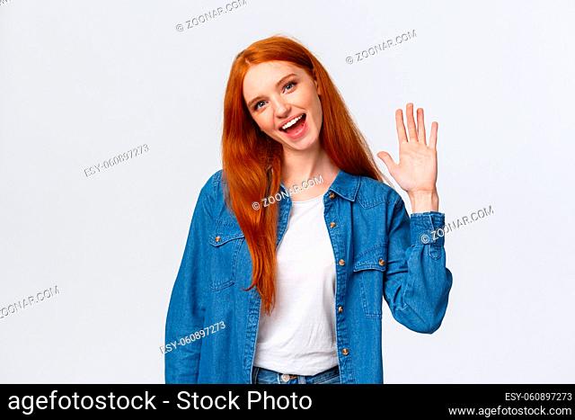 Friendly, cheerful and outgoing pretty redhead girl waving palm, saying hello, greeting team member, tilt head and smiling joyfully, nice meet you