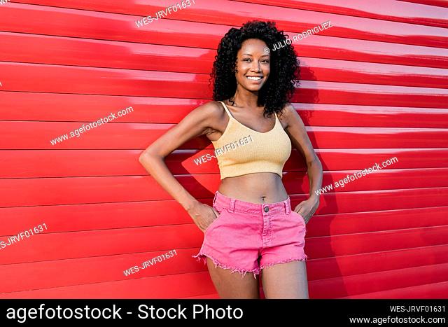 Smiling young woman standing with hands in pockets in front of red shutter