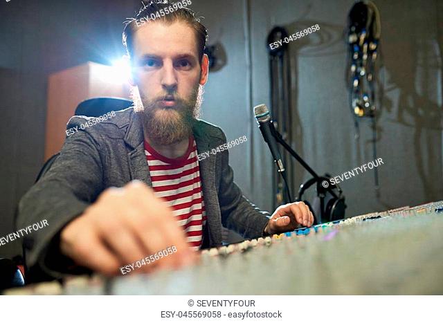 Young trendy man sitting at console in recording studio working and looking at camera