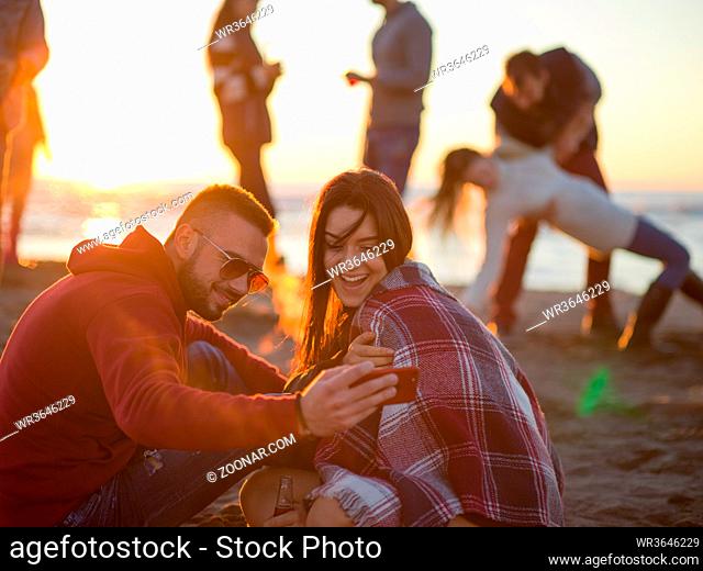 Couple using cell phone during autumn beach party with friends drinking beer and having fun